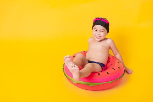 Summer vacation concept, Portrait Asian happy cute little child boy wear goggles, swimsuit hold beach watermelon inflatable ring, Kid have fun sit in inflatable, studio shot isolated yellow background