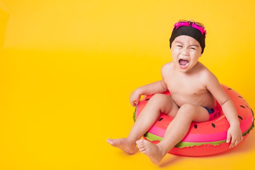 Summer vacation concept, Portrait Asian happy cute little child boy wear goggles, swimsuit hold beach watermelon inflatable ring, Kid have fun sit in inflatable, studio shot isolated yellow background