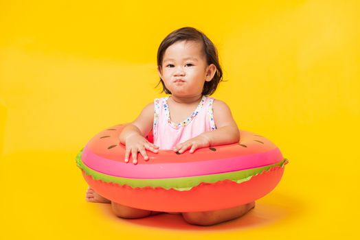 Summer vacation concept, Portrait Asian happy cute little baby girl wear swimsuit sitting in watermelon inflatable ring, Kid have fun sit in inflatable, studio shot isolated yellow background
