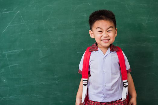 Back to School. Happy Asian funny cute little child boy from kindergarten in student uniform with school bag stand smiling on green school blackboard, First time to school education concept