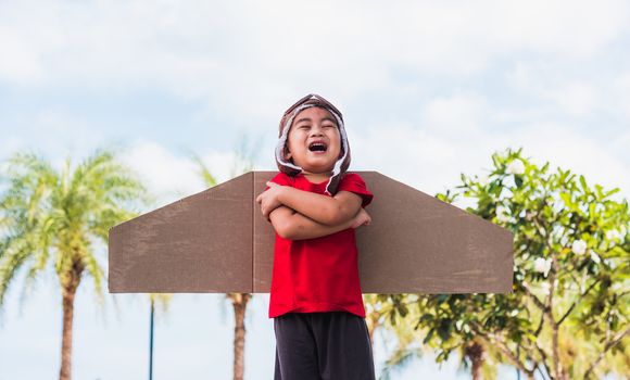Happy Asian funny child or kid little boy smile wear pilot hat and goggles play toy cardboard airplane wing fly stand crossed arm against summer sky cloud on garden background, Startup freedom concept