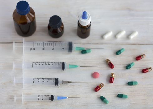 Syringes, capsules, pills and medical bottles lie horizontally on a white roughly painted wooden table.
