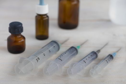 Medical syringes of various capacities and medical bottles lie diagonally on a white roughly painted wooden table.