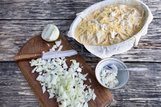 Ingredients for vegetable pie: chopped onions with knife, dough, feta, cheese on an old wooden weathered table.