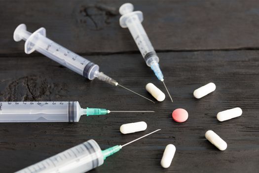 Medical syringes of various capacities, capsules, tablets lie on an old black wooden board and point to a red round tablet.