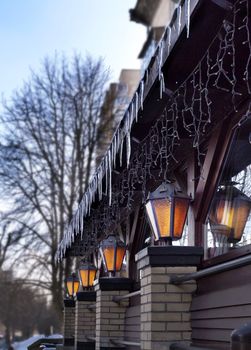Four old street lamps shine near the restaurant windows in the open air in the early spring morning. Icy icicles look down on them.