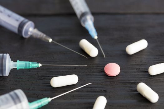 Medical syringes of various capacities, capsules, tablets lie on an old black wooden board and point to a red round tablet.