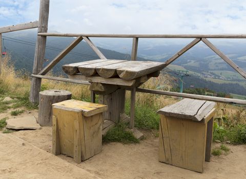 Carpathians. An old wooden weather table and three rough improvised chairs for a respite on top of the mountain. Ski lift in the background of a mountain landscape.
