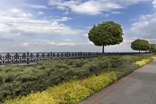 A paved stepped road in a beautiful park passes along the embankment of the river with ornamental flowers, framed decorative fence with cut bushes and lanterns for lighting.