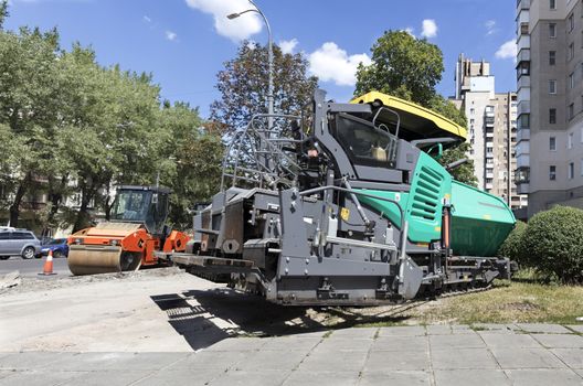 Among the summer noon, a heavy asphalt paver and road vibrating roller compactor is ready to repair a road in a modern city