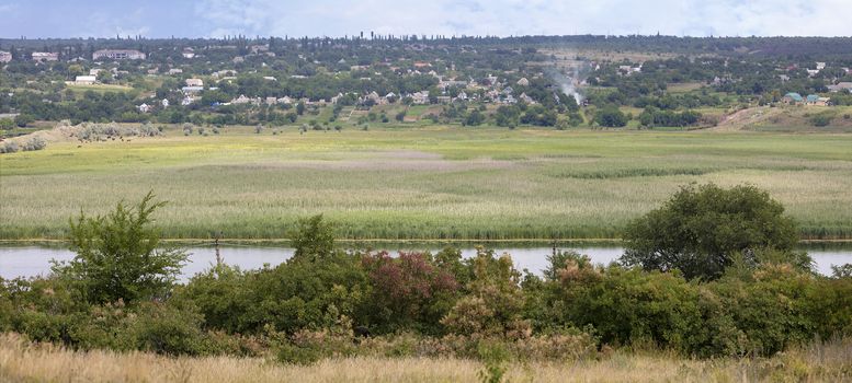 Scenic view from the opposite bank of the river to a village, which is located along the river with water meadows.