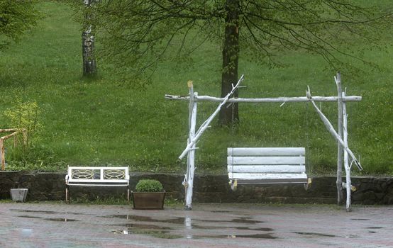 White swings in the garden after the summer rain are shrouded in foggy fumes against the background of juicy green grass and puddles on the pavement tile floor