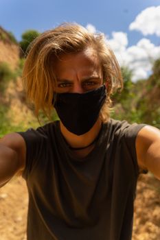 Self portrait of young man with medium long blond hair and blue eyes with mask in his mouth from the virus in the middle of the forest and nature