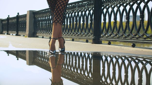 Female feet in high heels shoes walking in the puddle on the pavement. Slender beautiful woman legs reflecting in the water. Lady going in the city. Close up