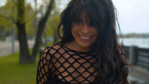 Close up portrait of attractive smiling woman in black clothes outdoors. She is standing on the embankment near the river in the city park. Real people