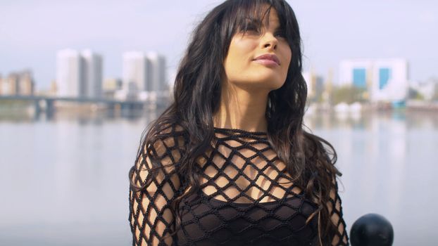 Close up portrait of a beautiful smiling dreamy woman on the background of the river. Woman in black dress posing on the embankment in the city park. Real people