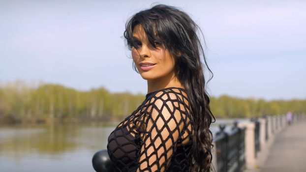 Close up woman with beautiful hair posing near the river. Portrait of smiling lady in black dress on the embankment in the city park. Real people