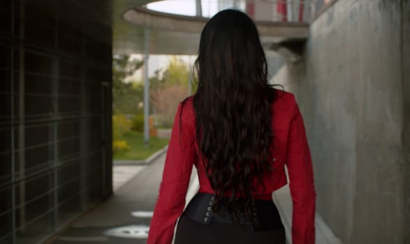 Rear view of attractive fashionable woman with long black hair in a red jacket walking in a city. Spring day, fresh leaves on the trees. Real people