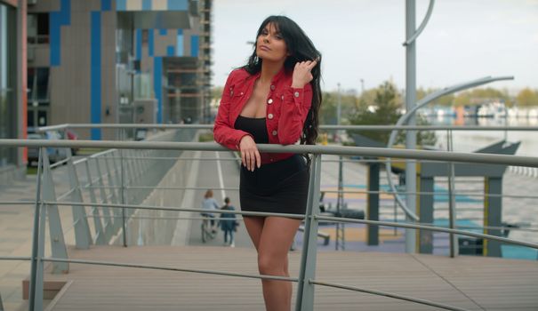 Calm woman with long beautiful hair standing on a bridge next to a modern building. Portrait of attractive girl in red jacket in the city. Real people