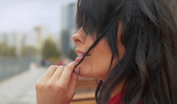 Close up profile portrait of beautiful woman with black long hair outdoors. She plays with a hair. Real people in the city.