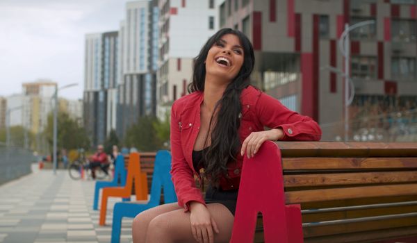 Close up portrait of beautiful brunette laughing woman in red jacket sitting on the bench outdoors. Real people in the city.