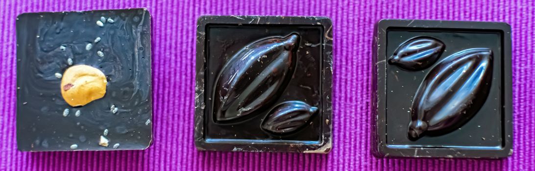 Handmade chocolate pieces with nuts are arranged in a row on a lilac background, close-up, selective focus. Useful and delicious sweets.