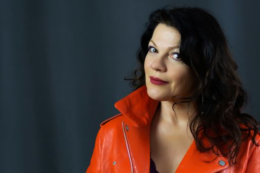 Portrait of a beautiful fashionable middle-aged woman in a bright orange leather jacket, posing on a dark gray background