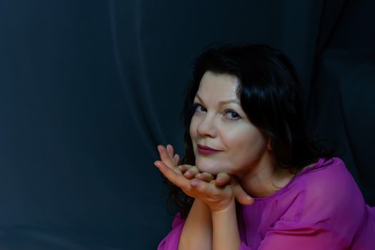 Portrait of a beautiful fashionable middle-aged woman in a bright pink semi-transparent long dress, posing on a dark gray background. The concept of preserving youth at any age.