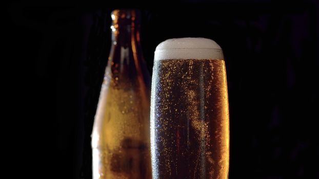 Close up full misted glass of beer on black background. Bubbles rising up and beatifully shining in the light beam. A bottle behind a glass