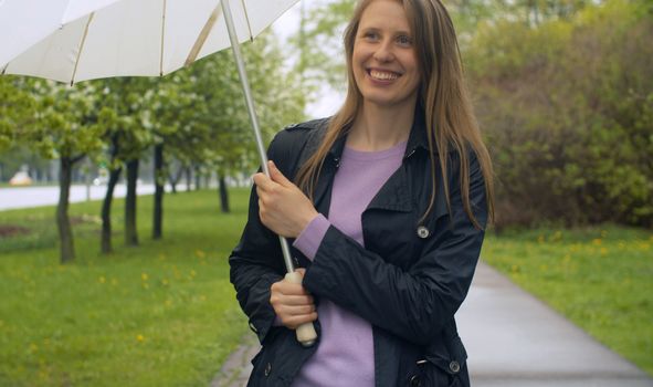 Rainy day in the park. Attractive smiling woman walking with umbrella under rain. Outdoors female portrait. Real people