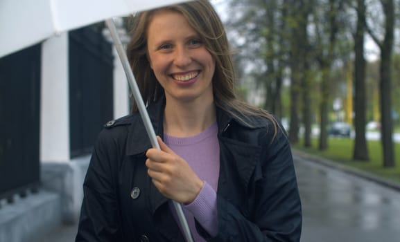 Spring rain in the city. Beautiful smiling woman with umbrella walking on the city sidewalk. Outdoors portrait of young blond woman. Real people.