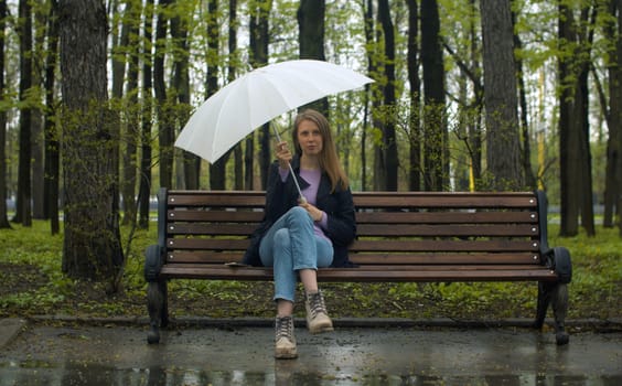 Portrait of a beautiful pensive woman with umbrella sitting on the bench in the park in spring. Rainy weather, real people