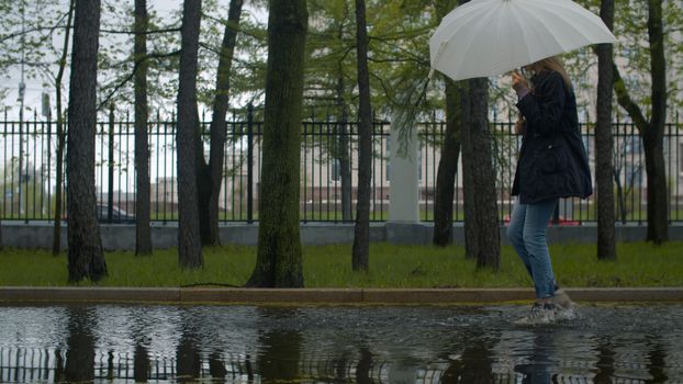 Cheerful woman with an umbrella in her hands walking in a puddle and spraying water with her feet. Rainy day in the park. Real people