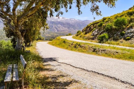 Two parallel mountain roads in Crete with a wooden bench in the foreground and snow-capped mountains in the background