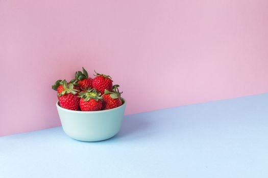 Minimalist image of a bowl full of strawberries on a light blue and pink background. Copy space available.