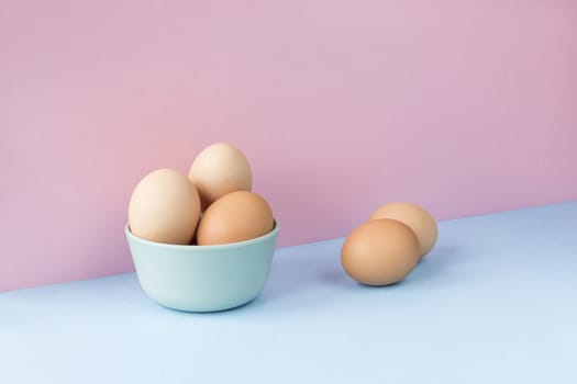 Minimalist image of several brown eggs in a bowl on a light blue and pink background. Copy space available.