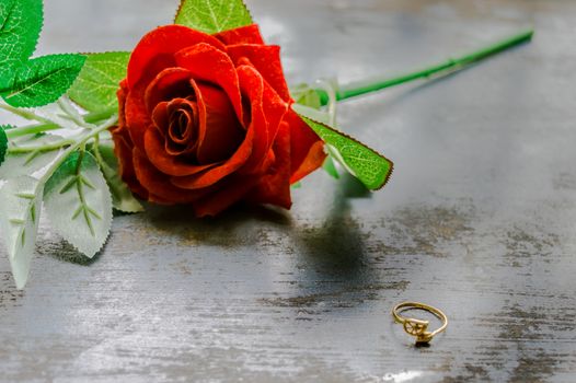 Close up gold engagement ring jewelery on rustic metal floor. Soft focus romantic Red rose flower in background. Love Proposal or Propose concept for valentines day wedding and holidays. Copy Space.