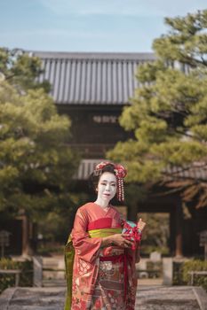 Maiko in a kimono posing in Kyoto on a stone bridge in front of the gate of a traditional Japanese kennin temple surrounded by pine trees.