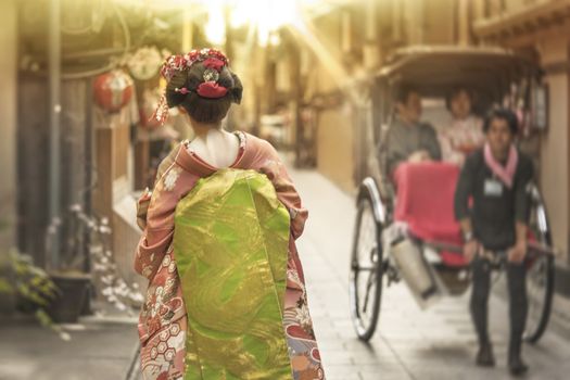 Maiko walking in an alley of Kyoto in the sunset light crossing a rickshaw.