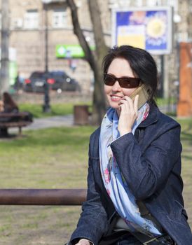 Portrait of a young, beautiful woman sitting on a bench in a spring park and smiling talking on the phone