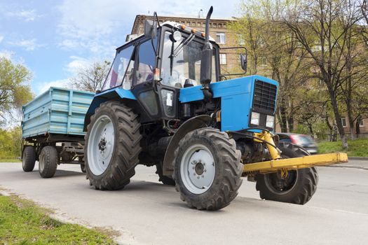 An old small blue tractor with a trailer stands on the edge of a city street road