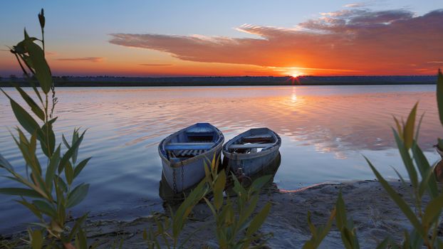 Two old wooden boats chained by the shore of a calm river. Early morning, the rays of the rising sun illuminate the summer landscape of the shore of a wide river.