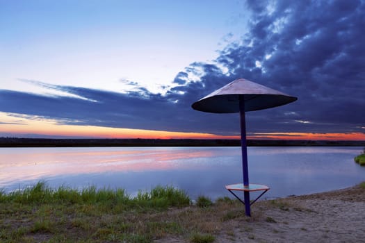 The bright rising sun illuminates the coastal grass, an old wooden canopy and bushes growing on the river bank
