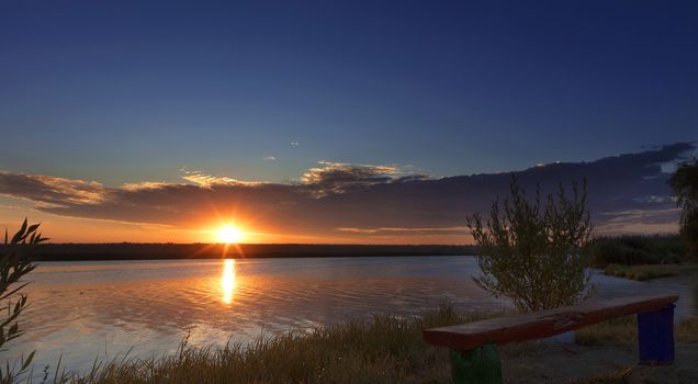 The bright rising sun illuminates the coastal grass, an old wooden bench and bushes growing on the river bank