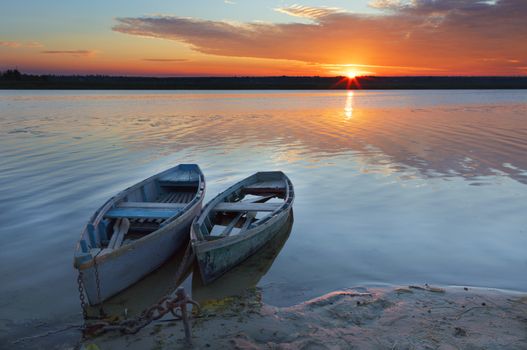 Two old blue-green wooden boats chained by the shore of a calm river. Early morning, the rays of the rising sun illuminate the summer landscape of the shore of a wide river.