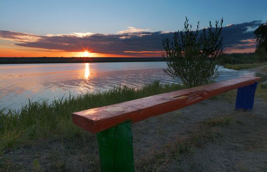 The bright rising sun illuminates the coastal grass, an old wooden bench and bushes growing on the river bank
