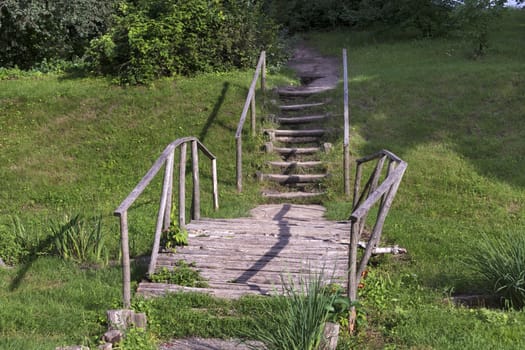 An old log wooden bridge over a stream with wooden steps, handrails and a dirt path leading to a hill