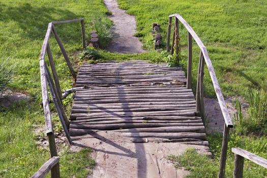 An old log wooden bridge over a stream with wooden steps, handrails and a dirt path leading to a hill