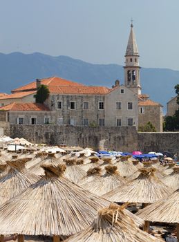 Thatched roofs of beach umbrellas in the bright sun with a defocused view of the tower of the old town of Budva
