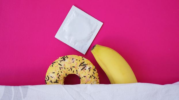 Sweet donut and banana on pink color background. Sex and Erotic concept. Banana hugs donut in bed. Condom - concept of protected sex